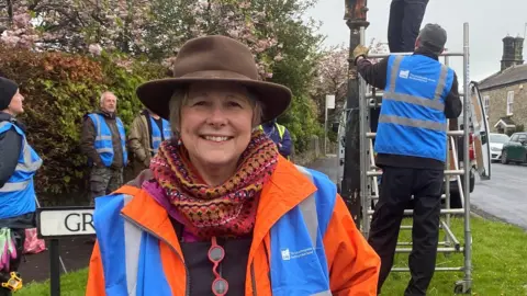 Annie Lloyd is a woman in her 50s with a warm smile and short brown hair. She is wearing a brown hat with a full brim, a pink patterned scarf and a blue and orange hi-vis jacket. Behind her a group is gathered around some scaffolding next to a fingerpost.
