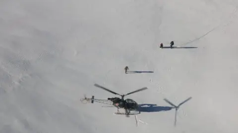 Reuters An aerial shot of a helicopter having landed on snow near two people with a third person walking between them.