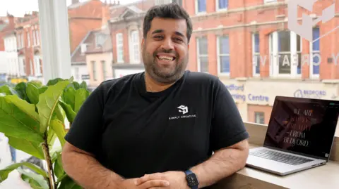  James Mall/Simply Creative Agency. Wasem Sebi, in a black top, smiling at the camera, standing by a laptop in his business, next to a green pot plant, on Bedford High Street