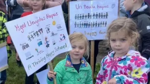 A child holds a protest banner which says 'Ysgol Llewyrchus Hapus'/'A thriving happy school'. They are standing next to another child and two other children can be seen behind them, along with a banner saying 'Un teulu hapus'/'One happy family'.