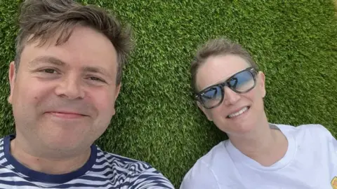 Geraint John Geraint and Deb lying on grass looking into the camera. He is wearing a navy and white striped t-shit and has short brown hair. She has her blonde hair tied back and is wearing sunglasses and a white t-shirt.