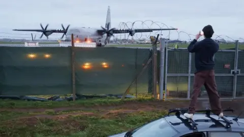 Reuters A man stands on the roof of a car in order to see over the perimeter fence of RAF Fairford. Temporary screening has been placed along the fence to try and discourage people from coming to watch activity inside. 