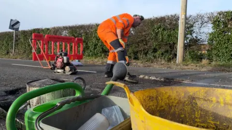 Road maintenance worker in orange jacket filling in a pothole on a road next to a hedge