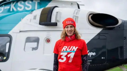 Air Ambulance Charity Kent Surrey Sussex A woman wearing an orange beanie and an orange t-shirt and long-sleeved shirt. She is standing in front of a helicopter.