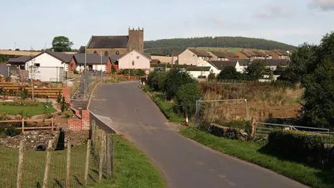 Walter Baxter The road leading to the village of Brydekirk with houses and a church clearly visible and a tree-lined hill in the distance