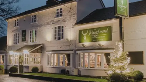 The front of a large white-walled brick pub in the evening, with lights illuminating the wall and a large green sign which reads The Haughmond