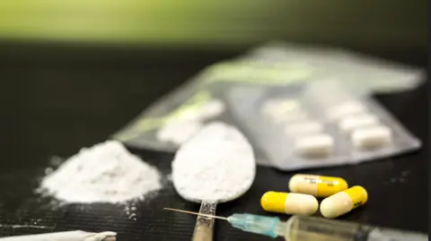 Getty Images White powder on spoon, alongside yellow and white pills and a needle, packets of pills in the background