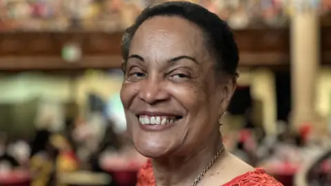Antonette, a lady in her 80s, smiles in a busy room, which is blurred behind her. She is wearing a gold necklace and a red top, and her black hair is back.