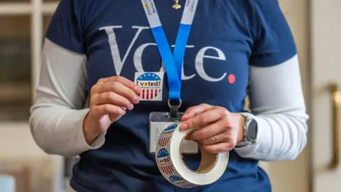 Kansas City Star via Getty Images A US poll worker wearing a VOTE T-shirt holds a roll of "I voted!" stickers