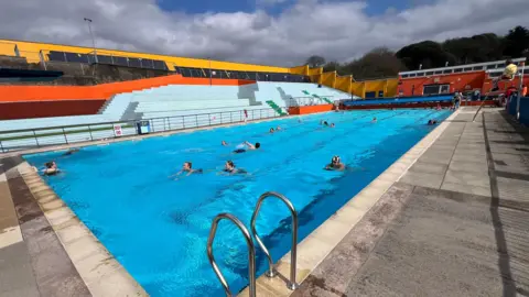 A view of the pool with dozens of swimmers in it. Coloured seating for the pool in the background, painted in bright orange, yellow and light blue.