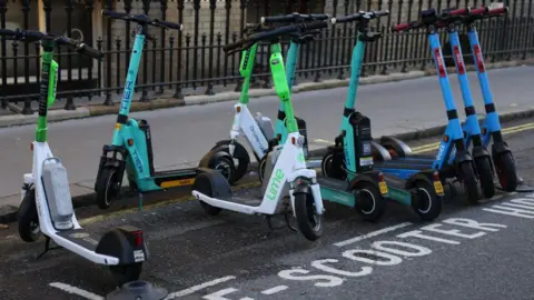 A row of electric scooters from various brands, including Lime, TIER, and Dott, are parked neatly in a designated "E-scooter hire" zone on the side of a city street. The scooters are lined up beside a black iron fence and a pavement.