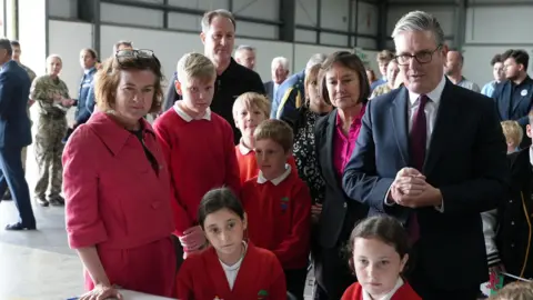 Eluned Morgan, Welsh Secretary Jo Stevens and Sir Keir Starmer stood around a group of children looking directly ahead. They are in a aircraft hanger and are listening to someone who is not in the shot.