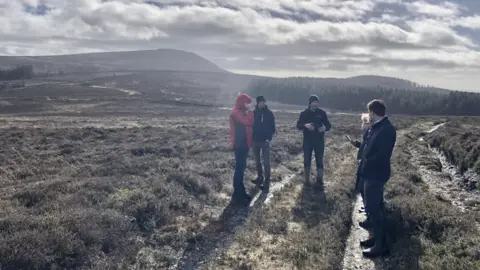 David Bellamy wears a beanie and waterproof coat as he talks to 4 people, they are stood on the reserve which is muddy and grassy, with hills in the background.