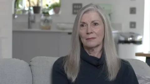 Carolyn Woods is sitting on a light grey sofa. She is looking sideways away from the camera and is wearing a black top. She has long fair hair and behind her, but not in focus, is a kitchen area