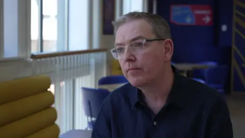 A man with grey hair and glasses is wearing a dark navy button-up shirt. He is sitting down and there are blue chairs in the background and a long, yellow sofa to his left