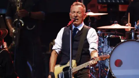 Bruce Springsteen performs on a stage. He is singing into a microphone and holding a guitar. A drum kit and saxophonist can be seen behind him. 