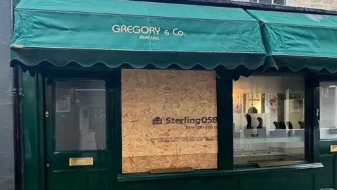 General street view of the boarded up shop front of jewelry store Gregory & Co. The window frames are painted green and there is a green marquis with the name written on it in gold letters. The large left-hand side window has been replaced by an OSB board which has 'Sterling OSB' written on it in bold black letters.