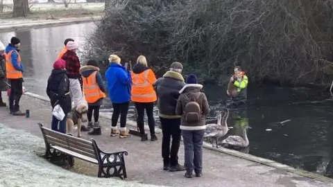 Oxford City Council Small crowd of people standing on a path looking towards a pond where a man is wading back to the shore holding a golden dog - the ground is white with ice.