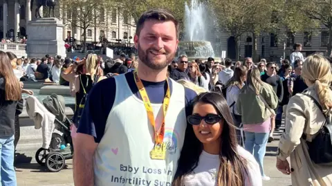 A man with a brown beard and short brown hair stands with his arm around a woman with long brown hair and sunglasses. The man wears activewear, a race number and a medal and the woman wears blue jeans and a white T-shirt. Both have text written across their shirts which reads "baby loss and infertility support".