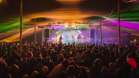 Chai Wallahs A festival scene inside a stretch tent lit up with green and purple lighting and a band on stage performing under a banner that says "Chai Wallahs". The tent is full of people and their backs are in view.