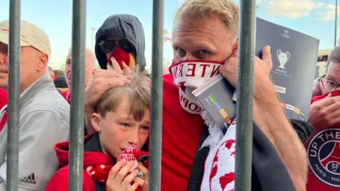 A man stands with his arms around a young boy who looks distressed. The man holds a Liverpool Football scarf around his nose and mouth and they are surrounded by other fans. They are standing behind metal bars. 
