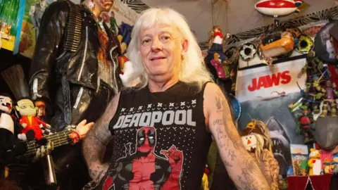 Martin leans against a red pool table surrounded by a dense collection of pop-culture memorabilia. The room contains masks, stuffed toys, posters, model displays and a life-sized figure dressed in black standing behind them.