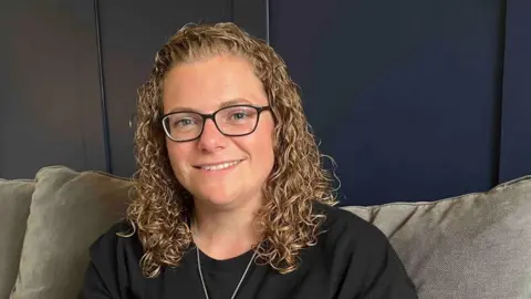 Aimee Tilley looking at the camera and smiling. She is wearing a black T-shirt and sitting on a grey sofa