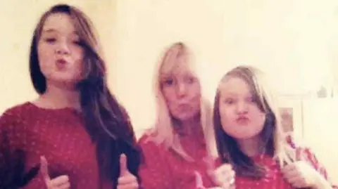 Family photo Two young girls with long brown hair, with a woman with long blonde hair in between them. They are all wearing matching red pyjamas, pulling pouting faces and have their thumbs up as they look at the camera.