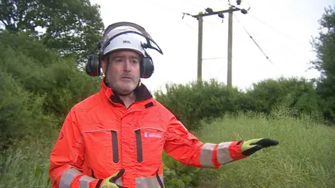 A worker in an orange jacket stands next to overhead lines in the middle of the countryside