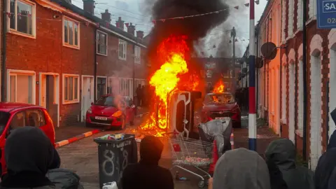PA Media An overturned vehicle burns in the middle of a residential street as thick black smoke rises into the sky. Flames have spread to a nearby car, while a crowd of hooded onlookers watches from behind makeshift debris on the road. Union flags hang overhead between rows of terraced houses.