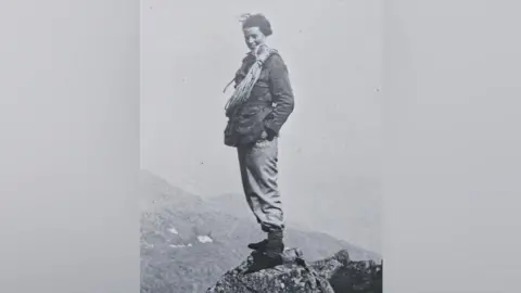 Mountain Heritage Trust Alice 'Jammy' Cross atop Scafell Pike in 1939. A black and white photograph (in the early days of cameras) shows Alice with a big smile on her face and a large rope resting on her shoulders. She is wearing a thick coat and wide trousers and has them tucked into her socks.