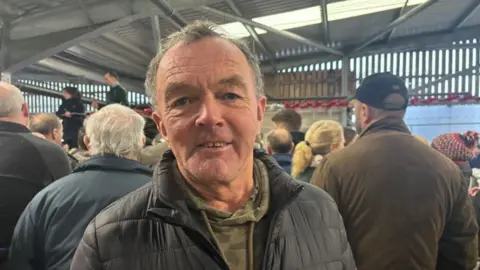 ALEX JAMES/BBC Robin pictured looking into the camera and smiling slightly. He's wearing a camouflage green hoodie and a black puffer jacket. He's stood in the midst of a crowd of people inside the cattle market - which is a large warehouse or barn-type structure. 