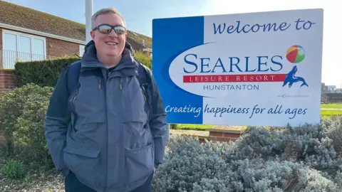 Richard Brown is a holiday maker at Searles holiday park. He is wearing sunglasses and standing in front of a sign welcoming people to stay at Searles holiday park in Hunstanton. 