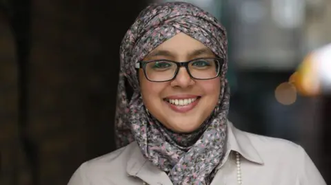 A woman in a floral headscarf and beige trench coat smiling at the camera