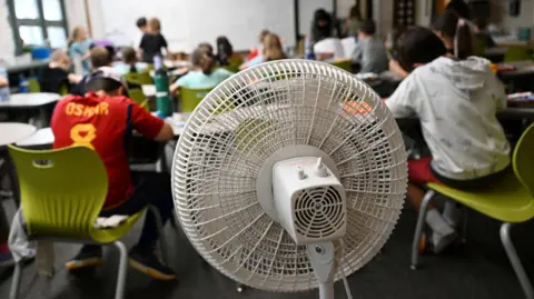 A picture taken from the back of a classroom showing a large white fan blowing air towards young children studying at their desks