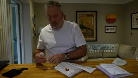 Andrew Parker preparing a stoma kit on a wooden table in his home. The small white packet is open and Andrew is placing items inside of it. He is wearing a white t-shirt and a watch. There is a sofa behind him and different framed artworks are hanging on the wall.
