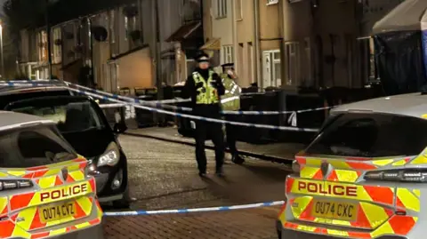 Shariqua Ahmed/BBC A street at night. There are two police cars in the foreground, and a couple of officers. There is blue and white police tape across the street.