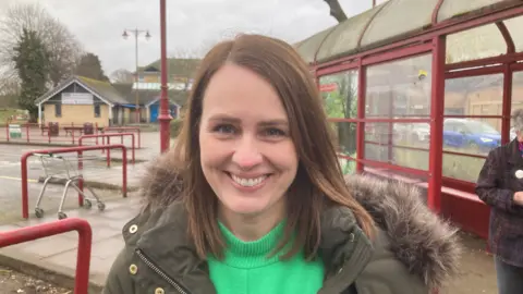 Martin Heath/BBC Hayley Charlesworth with long brown hair smiling at the camera. She is wearing a green coat with a furry hood and a light green top. There is a red-framed glass bus shelter behind her. There is a shopping trolley to the left.