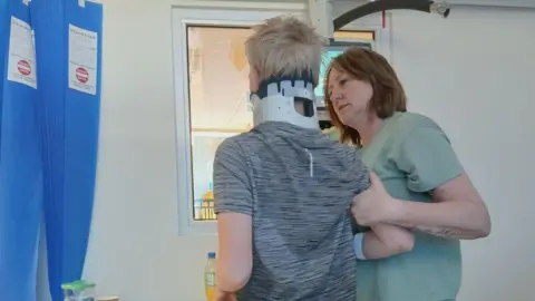 Jaime Owen A woman with shoulder length brown hair and wearing a green top, holds a 12-year-old boy by his right arm. The boy, who has his back to the camera, is in a hospital bay and is wearing a neck brace and a grey t-shirt. 
