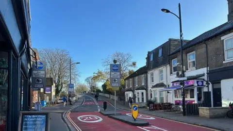 View of part of Mill Road in Cambridge. Road markings show it is a 20mph zone and there are red marking to indicate the bridge is a bus gate. Signs before the bridge show restrictions that are in place. There are shops and restaurants at the sides of the road