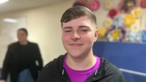 Riley is wearing a black Nike jacket and bright purple top. He is smiling at the camera and has brown hair.