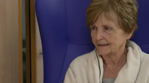 Female patient with dark blond hair wearing a cream dressing down and grey t-shirt. She is sitting on a large purple leather chair in a hospital ward.