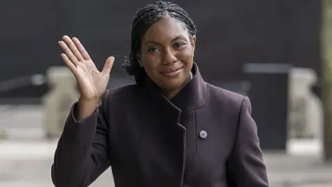 Getty Images Kemi Badenoch waving and smiling. She is wearing a brown jacket with a high neck collar. She has black hair.
