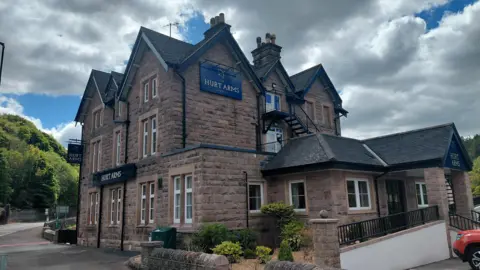 A general view of the pub from the road. The building is light stone coloured with a dark tiled roof.