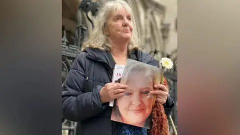 Joy Dove outside a court holding a picture of her smiling daughter. In her left hand she is holding a white flower. 