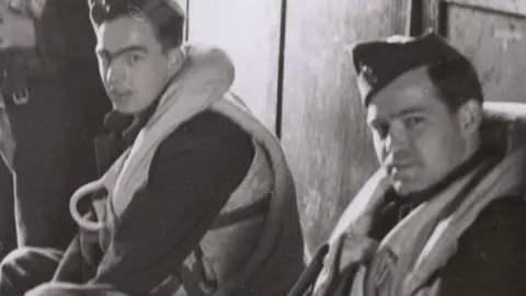 A black and white image showing a close up of two Lancaster pilots in a locker room at RAF Duxford during World War Two. The men are sitting down, in uniform with life guards and looking towards the camera.