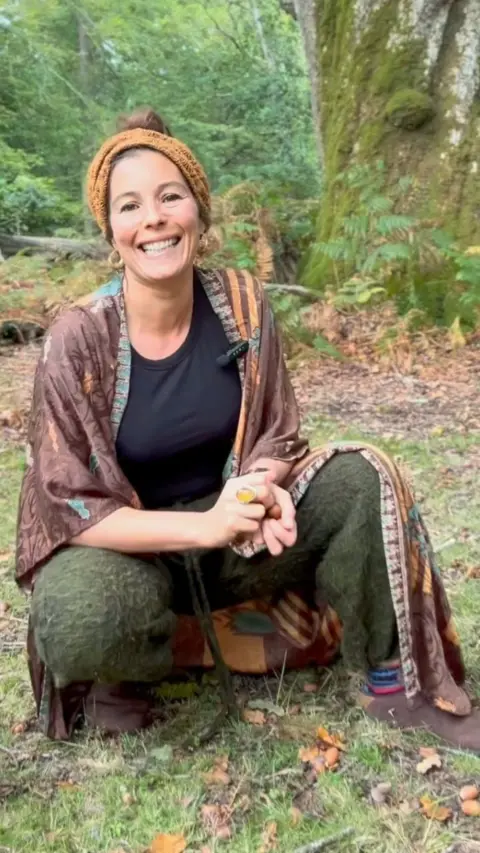 Sammie Longhorn crouches down in the grass in front of a tree. She is grinning as she poses for a photo with her hands clasped together. She has brown hair tied back in a bun with a dark yellow cloth headband and a brown and green kaftan.