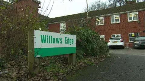 A sign that is half white and green has green writing saying 'Willows Edge'. It is about ankle height and is in front of a two-storey red-brick building.