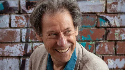 The Wallace Collection John Wallace is looking to the side of the camera and is smiling broadly. He has greying floppy hair and is wearing a blue shirt and a beige linen jacket. He is standing in front of a brick wall with graffiti on it