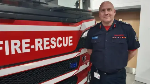 Simon is bald and is standing next to a large red and white fire engine that says "fire-rescue" on the front of it. He is wearing a dark navy shirt which says "fire and rescue" on the front